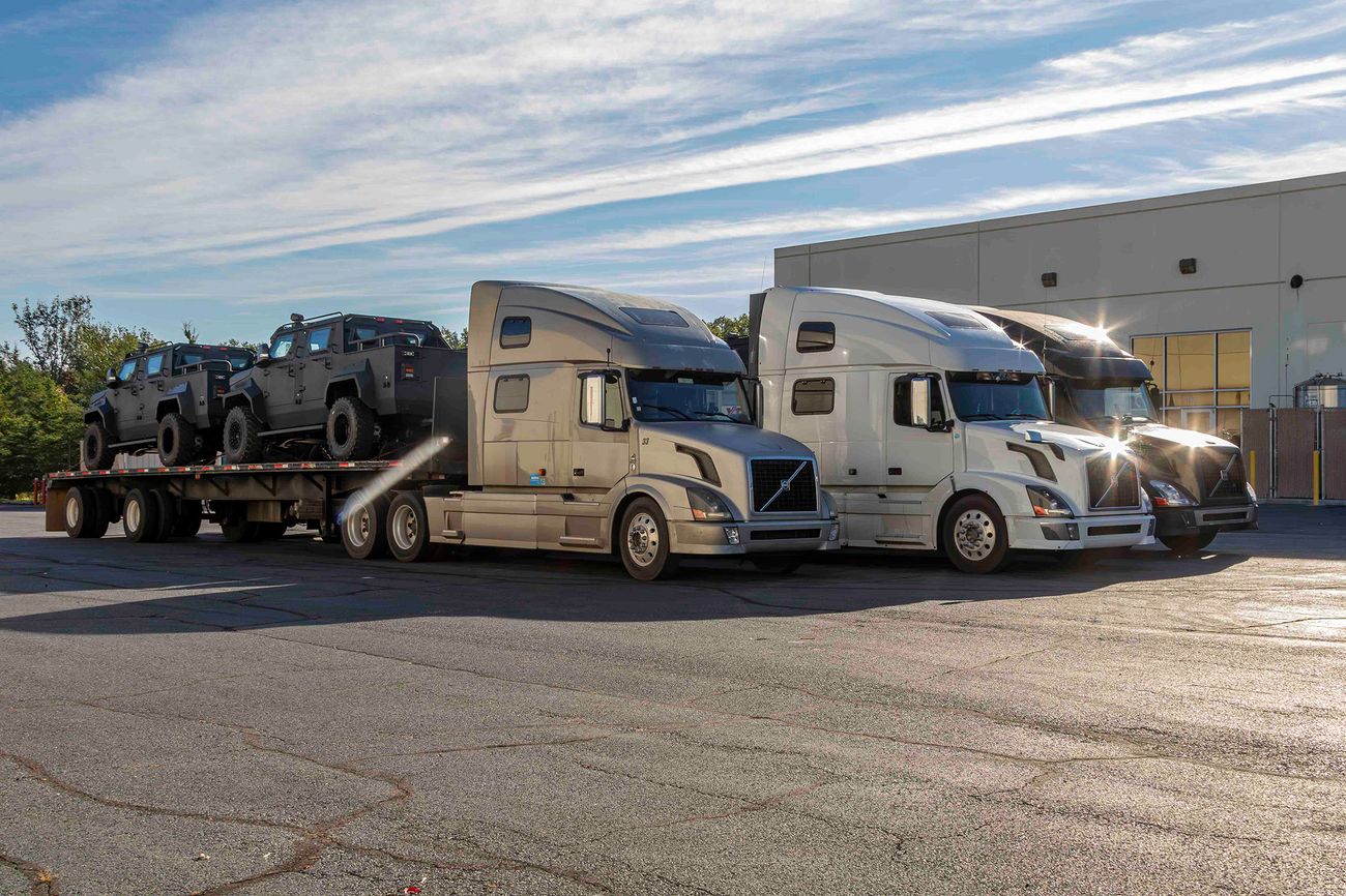 Three semi-trucks parked in front of a warehouse, one with a flatbed trailer carrying multiple black armored vehicles from Alpine Armoring.
