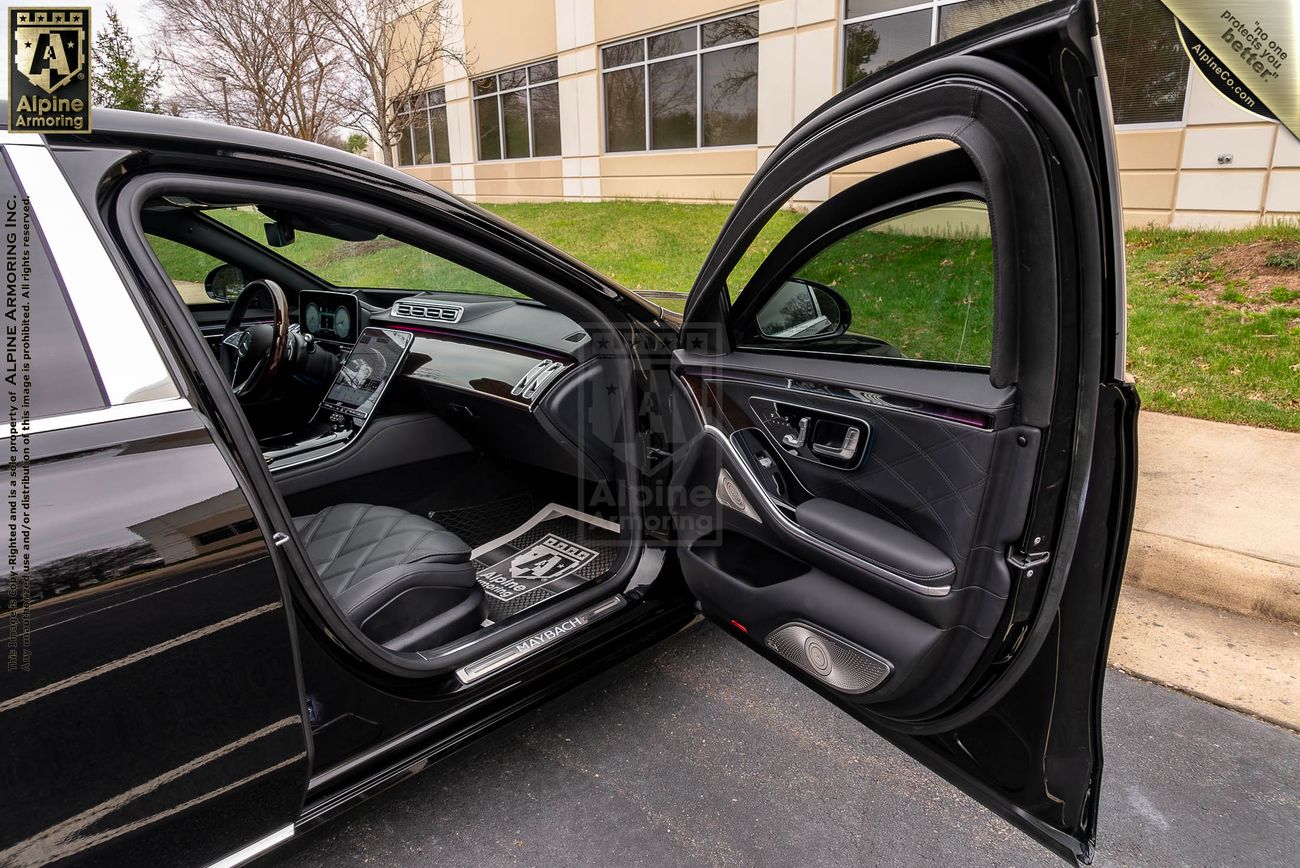 An open door of a black armored Mercedes-Benz S 580 Maybach sedan revealing the interior, including a plush seat and a mat on the floor. The vehicle is parked next to a building with bulletproof glass windows and a lawn area outside.