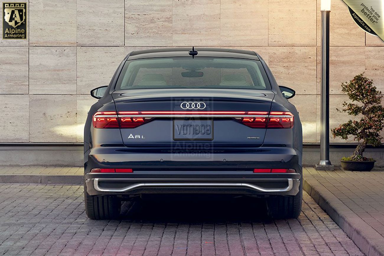 A rear view of a parked, dark-colored Audi A8L sedan against a stone wall backdrop, with an Alpine Armoring logo displayed on the top-left corner.