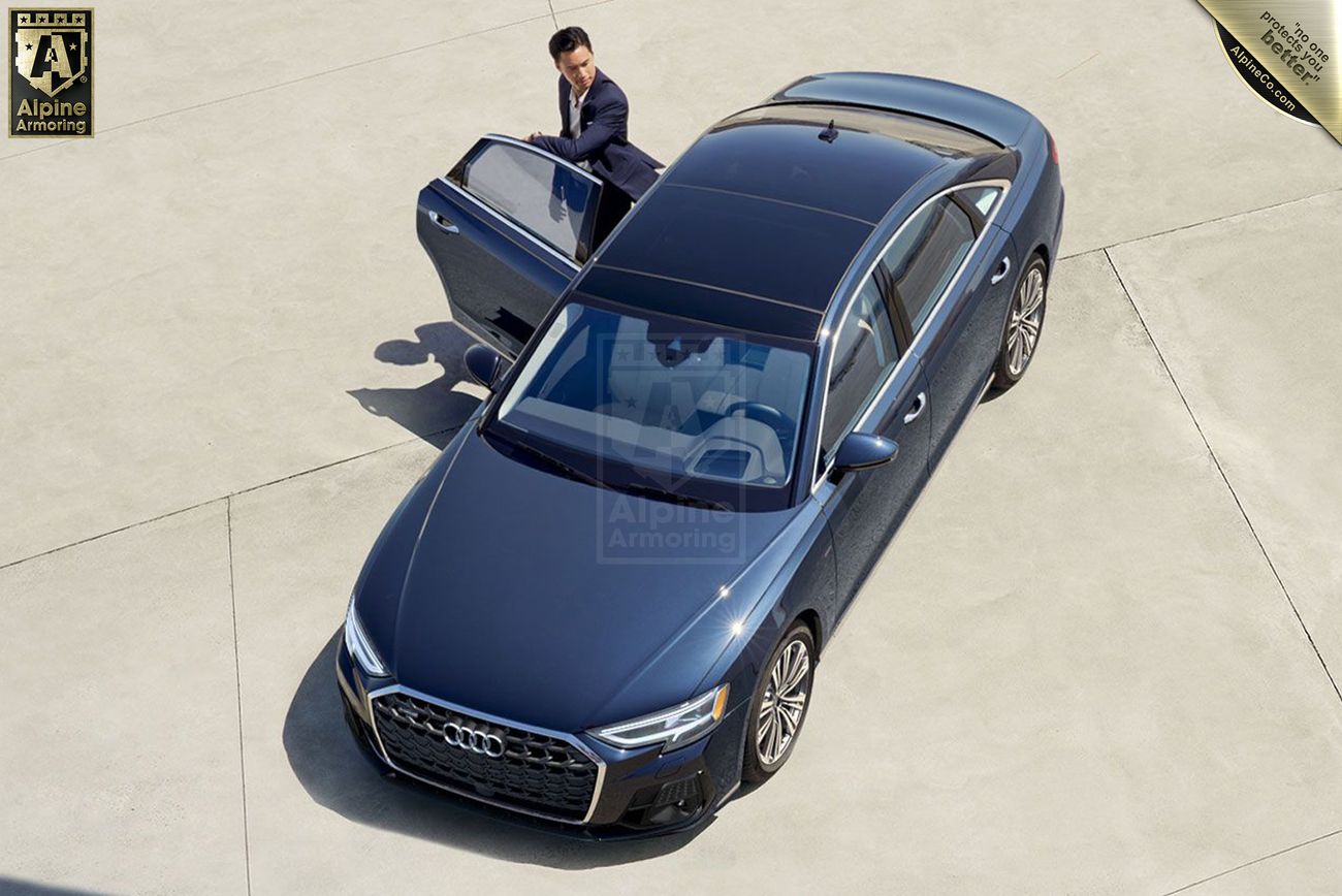 A person in a suit opens the driver's door of a dark blue Audi sedan parked on a concrete surface. An Alpine Armoring sign is visible in the background.