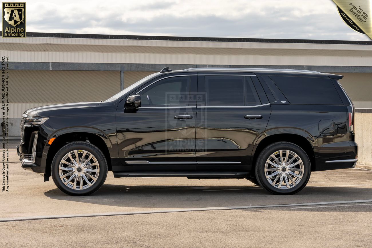 A black, full-size armored Cadillac Escalade SUV is parked on a paved area in front of a building under a cloudy sky. The vehicle is shown in a side view.