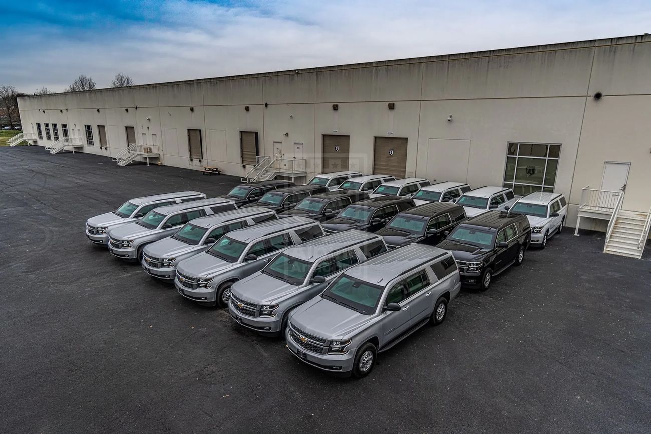 A line of white pickup trucks is parkednext to the Alpine Armoring Headquarters building on an overcast day.