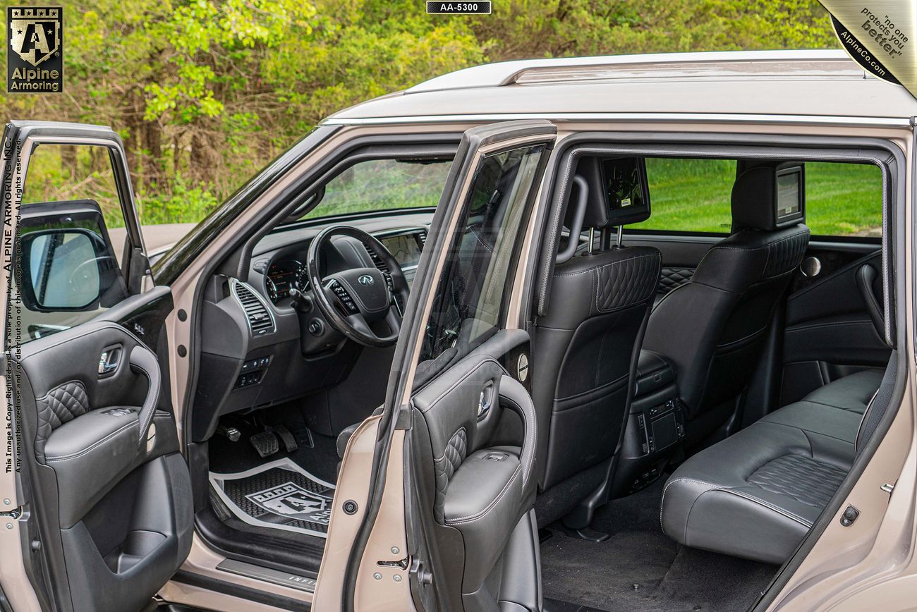 Interior view of a INFINITI QX80 SUV with open doors, showing black leather seats, a mounted screen on the back of the front seat, and an intricate floor mat design. The background features greenery.