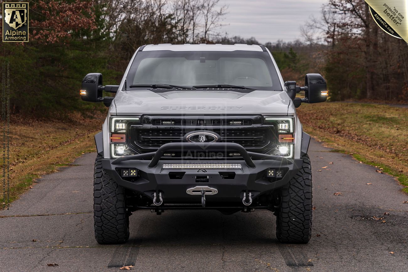 Front view of a white Mastiff truck with black accents, fitted with off-road tires, a spare tire on the rear, and a roll bar, parked on a road with trees and overcast sky in the background.