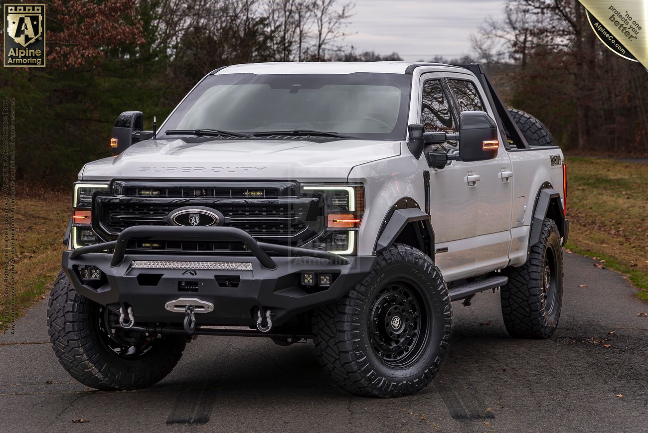 A white Mastiff truck is parked on a paved road surrounded by trees. The truck features large off-road tires, a front bumper with a winch, and a roof rack.