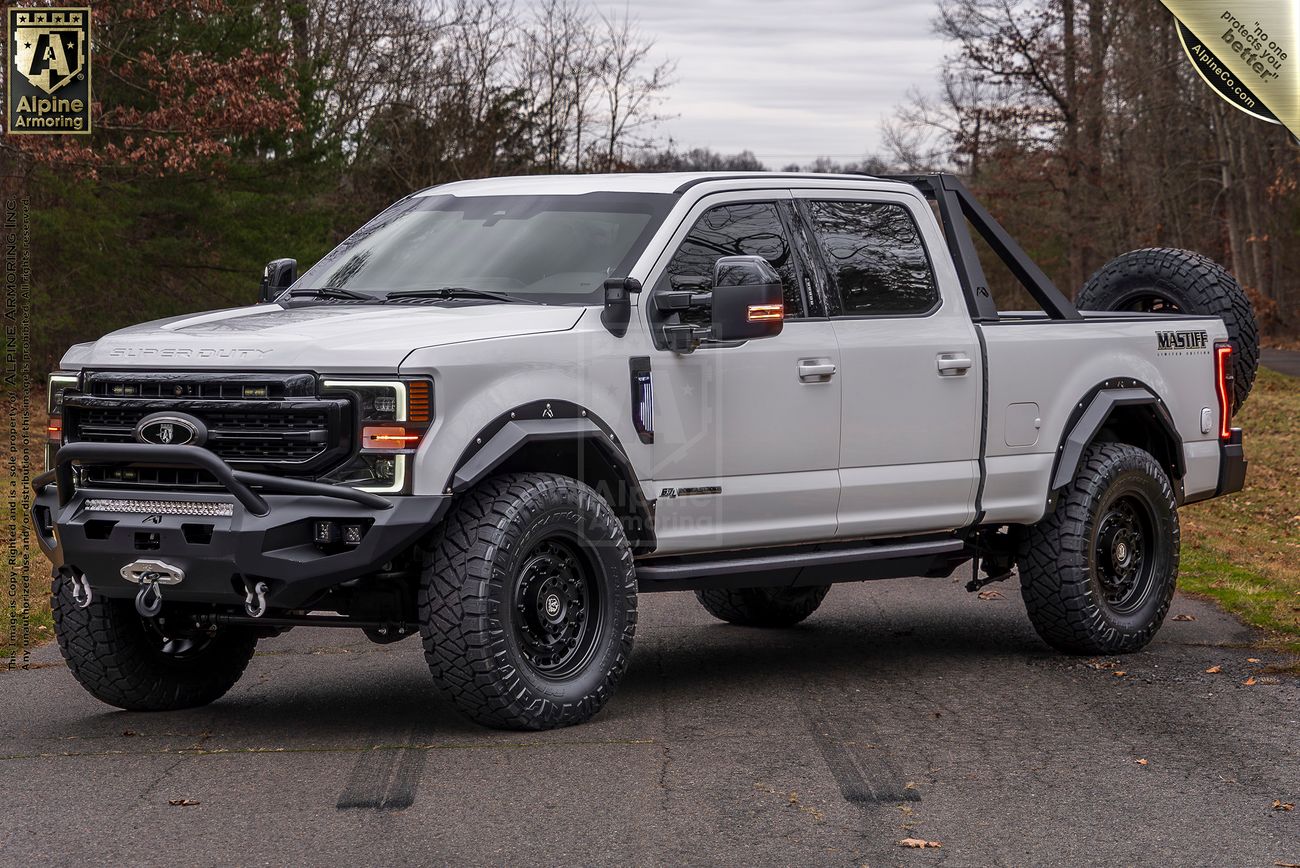 Front view of a white Mastiff truck with black accents, fitted with off-road tires, a spare tire on the rear, and a roll bar, parked on a road with trees and overcast sky in the background.