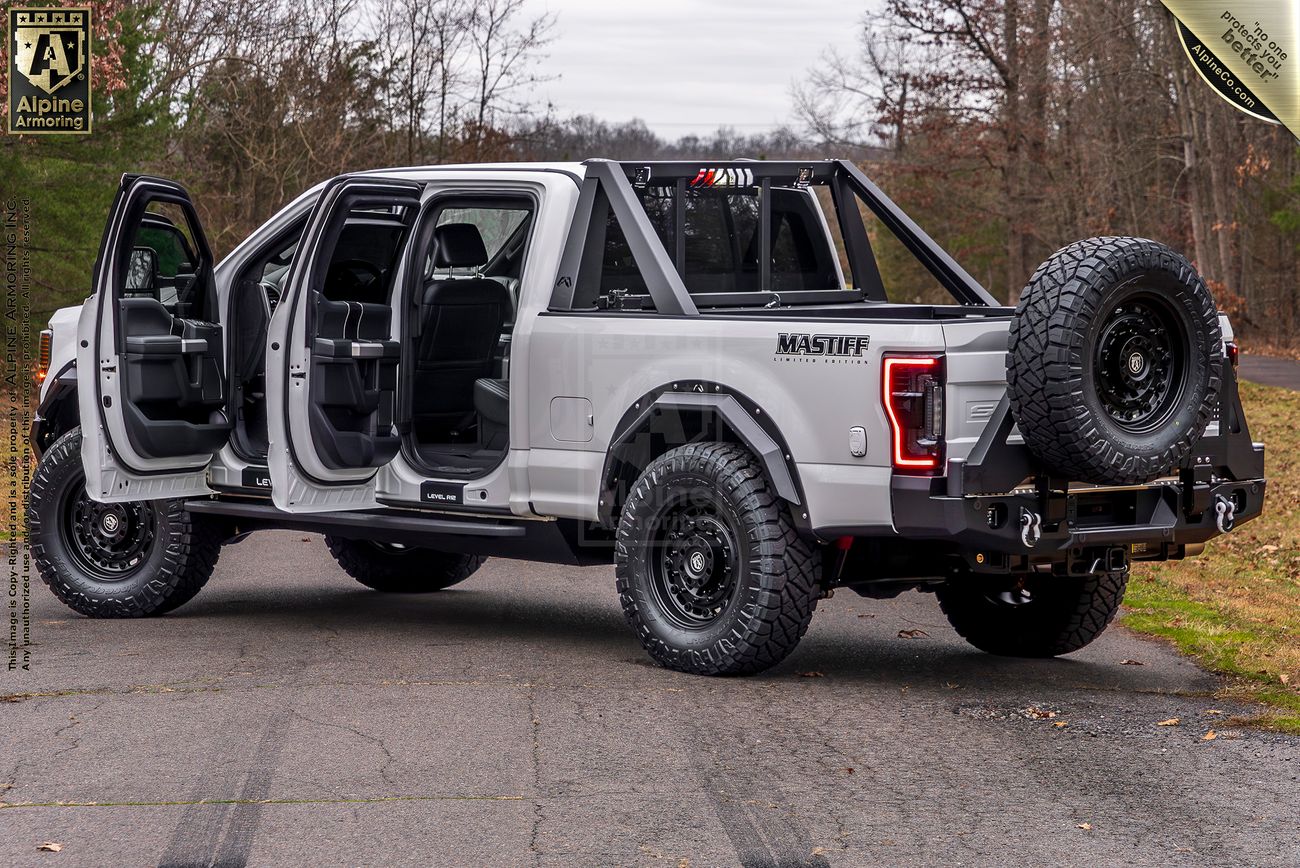 A silver customized Mastiff with off-road tires, open doors, and a spare tire mounted on the back, parked on a paved surface with trees in the background.