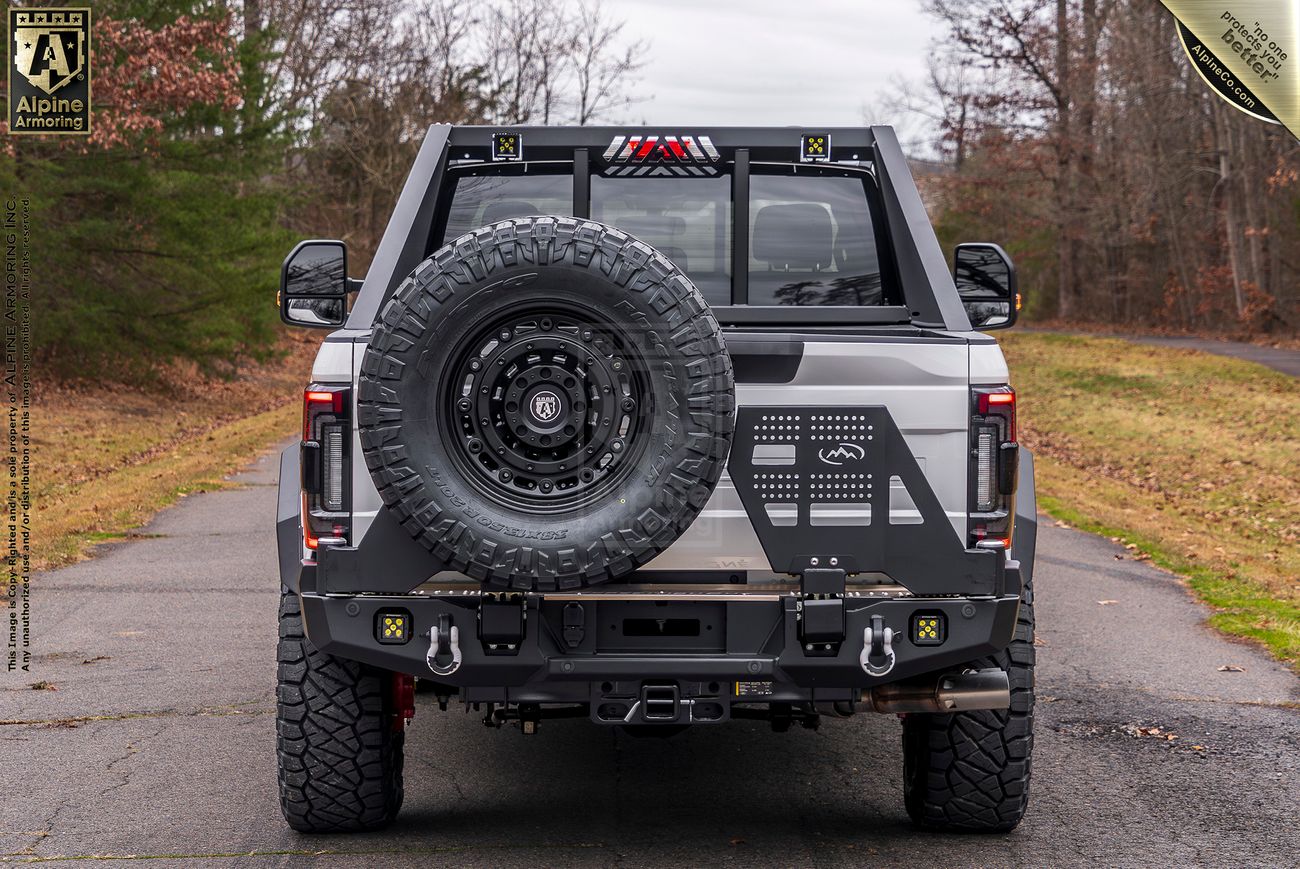 Rear view of a white Mastiff truck with black accents, fitted with off-road tires, a spare tire on the rear, and a roll bar, parked on a road with trees and overcast sky in the background.