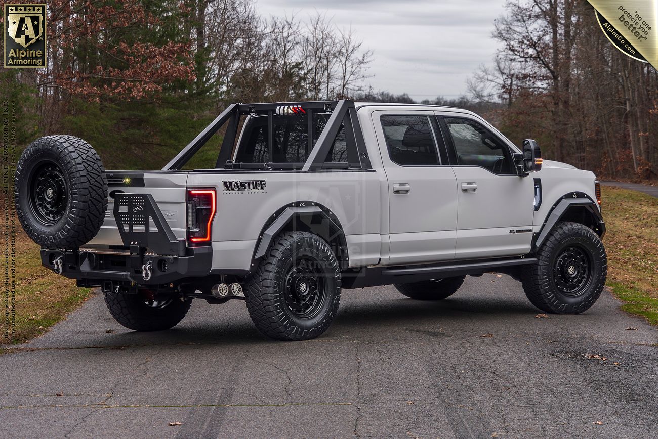 Rear view of a white Mastiff truck with black accents, fitted with off-road tires, a spare tire on the rear, and a roll bar, parked on a road with trees and overcast sky in the background.
