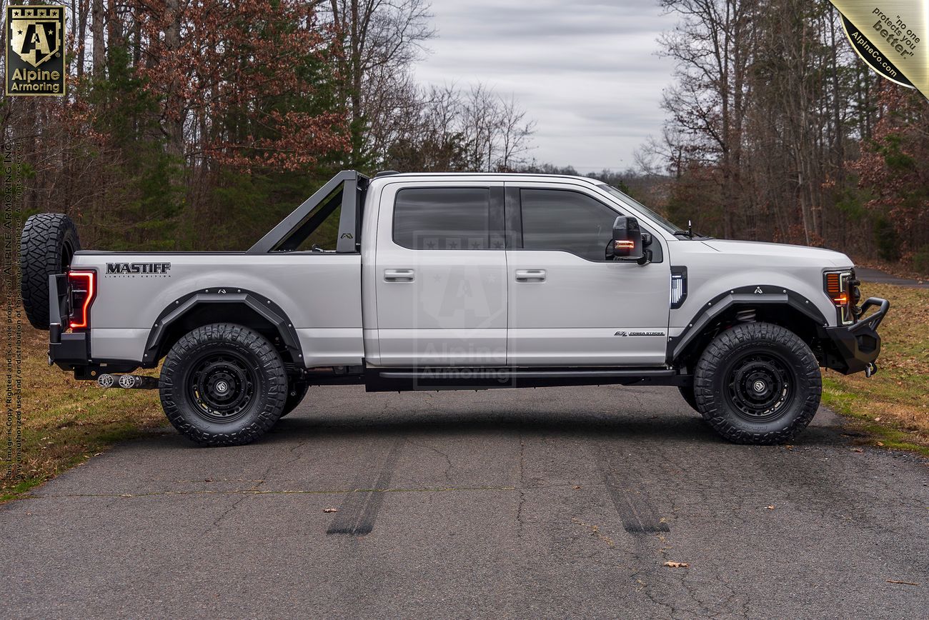 Side view of a white Mastiff truck with black accents, fitted with off-road tires, a spare tire on the rear, and a roll bar, parked on a road with trees and overcast sky in the background.