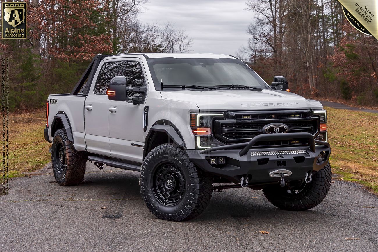Front view of a white Mastiff truck with black accents, fitted with off-road tires, a spare tire on the rear, and a roll bar, parked on a road with trees and overcast sky in the background.
