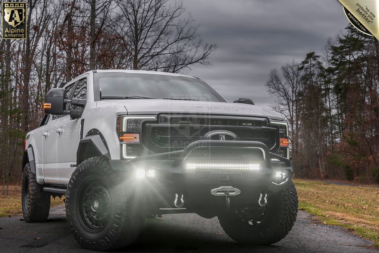 A white lifted Mastiff with off-road tires and LED lights drives on a paved road surrounded by leafless trees under a cloudy sky. 