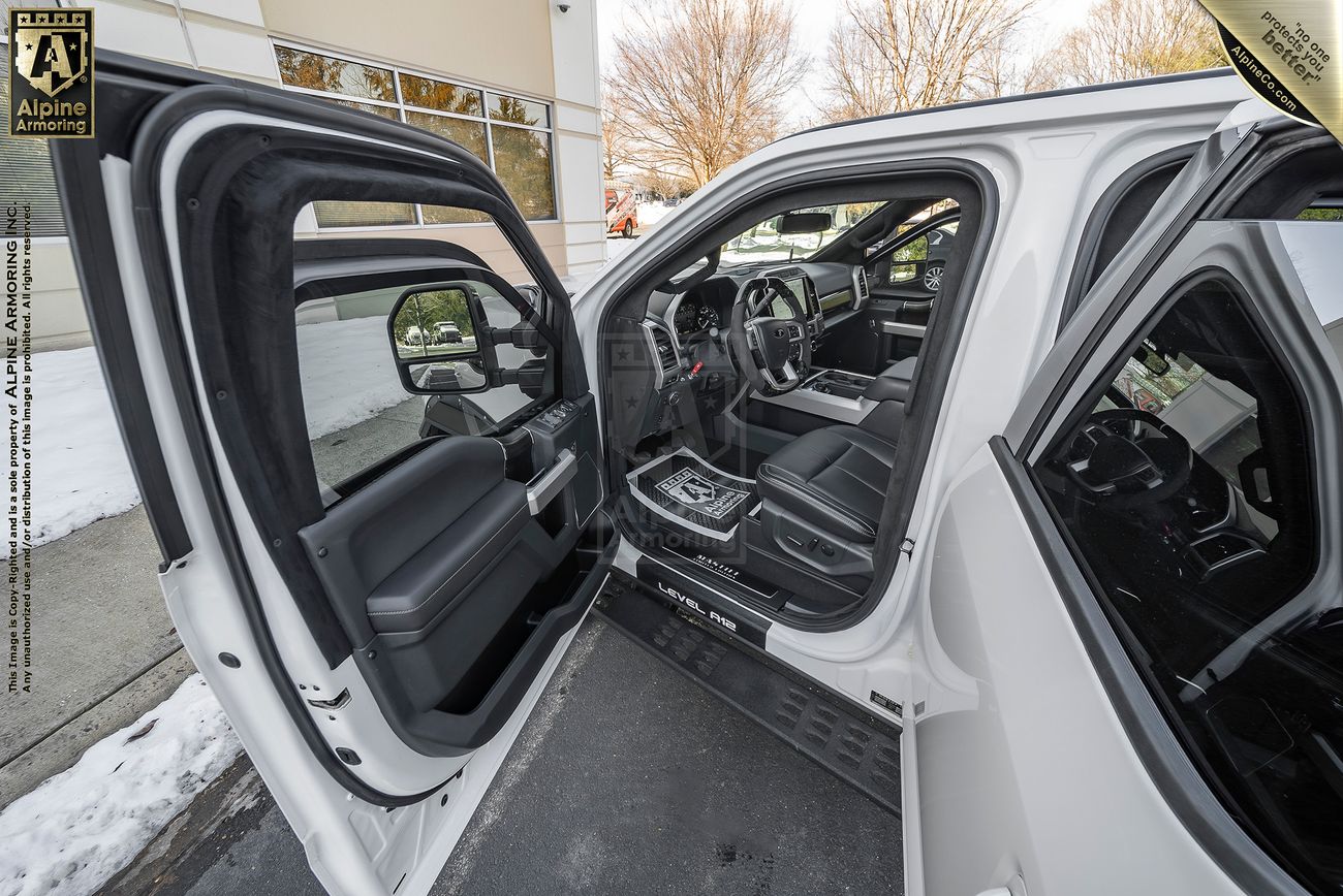 An open driver door shows the interior of a white Mastiff parked outdoors near a snowy sidewalk, with visible trees and a building exterior in the background.