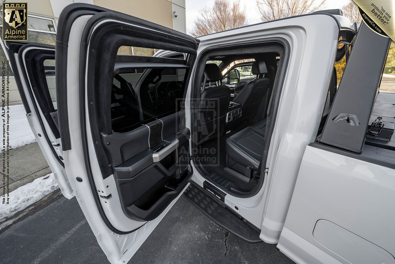 An open rear driver-side door shows the interior of a white Mastiff parked outdoors near a snowy sidewalk, with visible trees and a building exterior in the background.