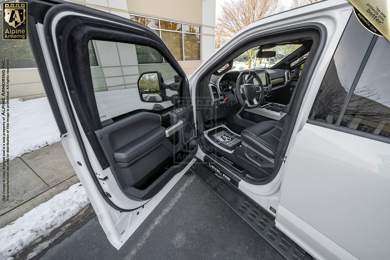 An open driver door shows the interior of a white Mastiff parked outdoors near a snowy sidewalk, with visible trees and a building exterior in the background.