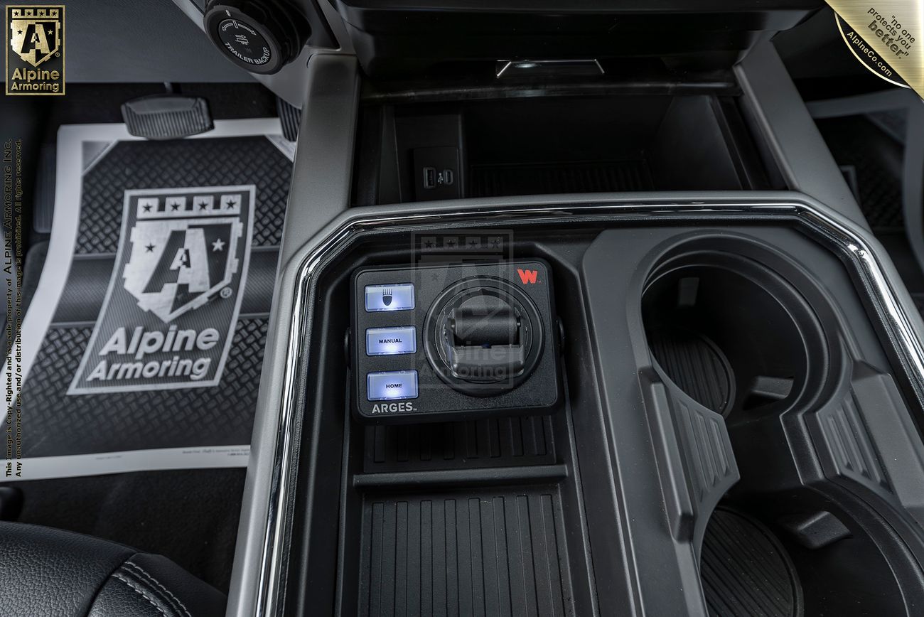 Close-up of a Mastiff's center console featuring cup holders, an Alpine Armoring floor mat, and a set of buttons with blue lights and a rotary knob labeled with a red "W".