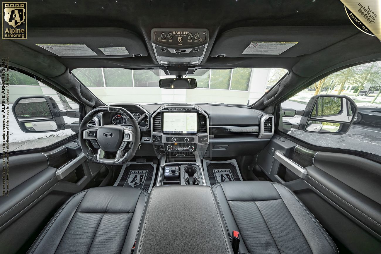Panoramic view of a Mastiff's interior, showing the steering wheel, dashboard, touchscreen display, and leather seats. The design is modern with metallic and leather accents. Alpine Armoring branding is visible.