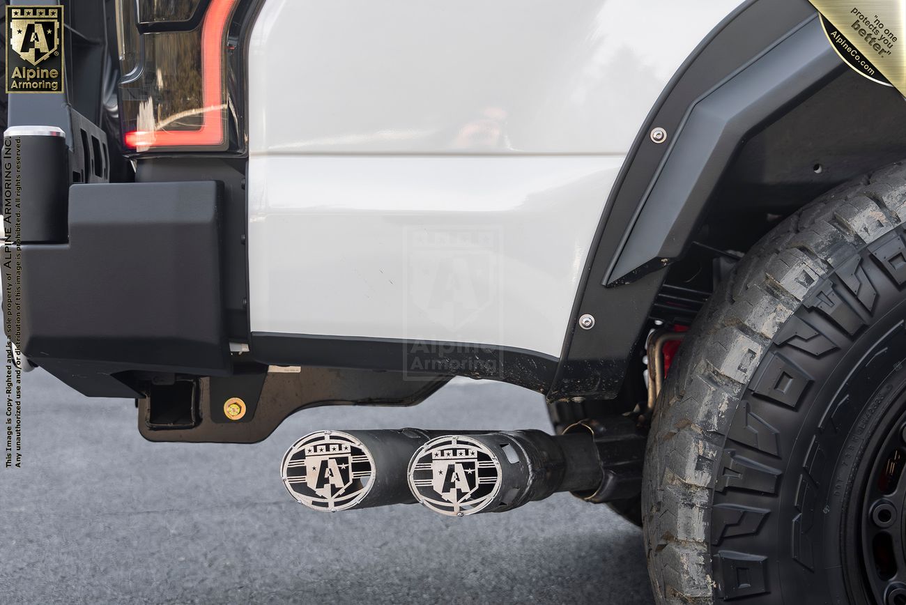 Close-up image of a Mastiff's rear, showing dual exhaust pipes with custom metallic covers featuring the Alpine Armoring logo, and part of a rugged off-road tire.