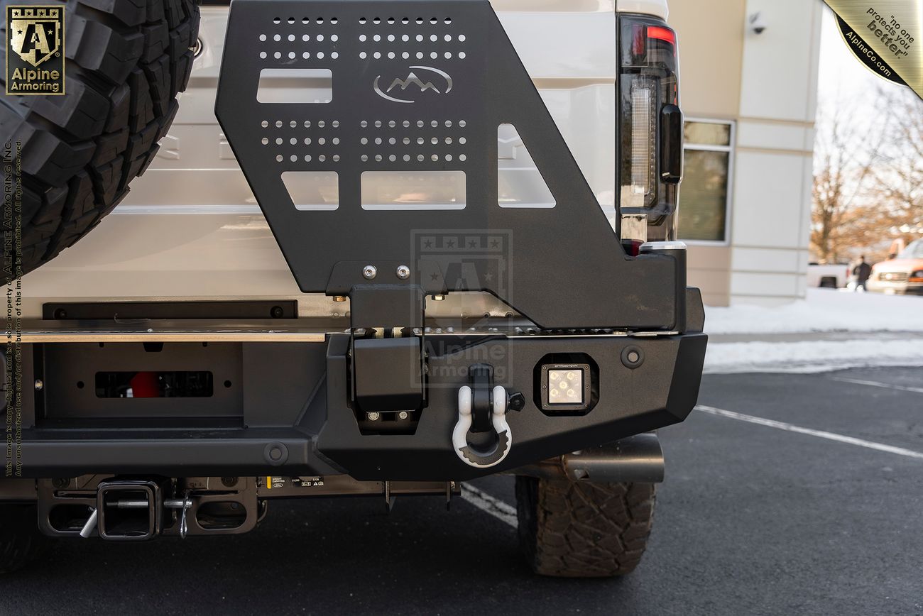Close-up of the rear bumper of a white Mastiff with a custom black metal plate, tow hook, and off-road tire. Snow is visible on the ground near a building in the background.
