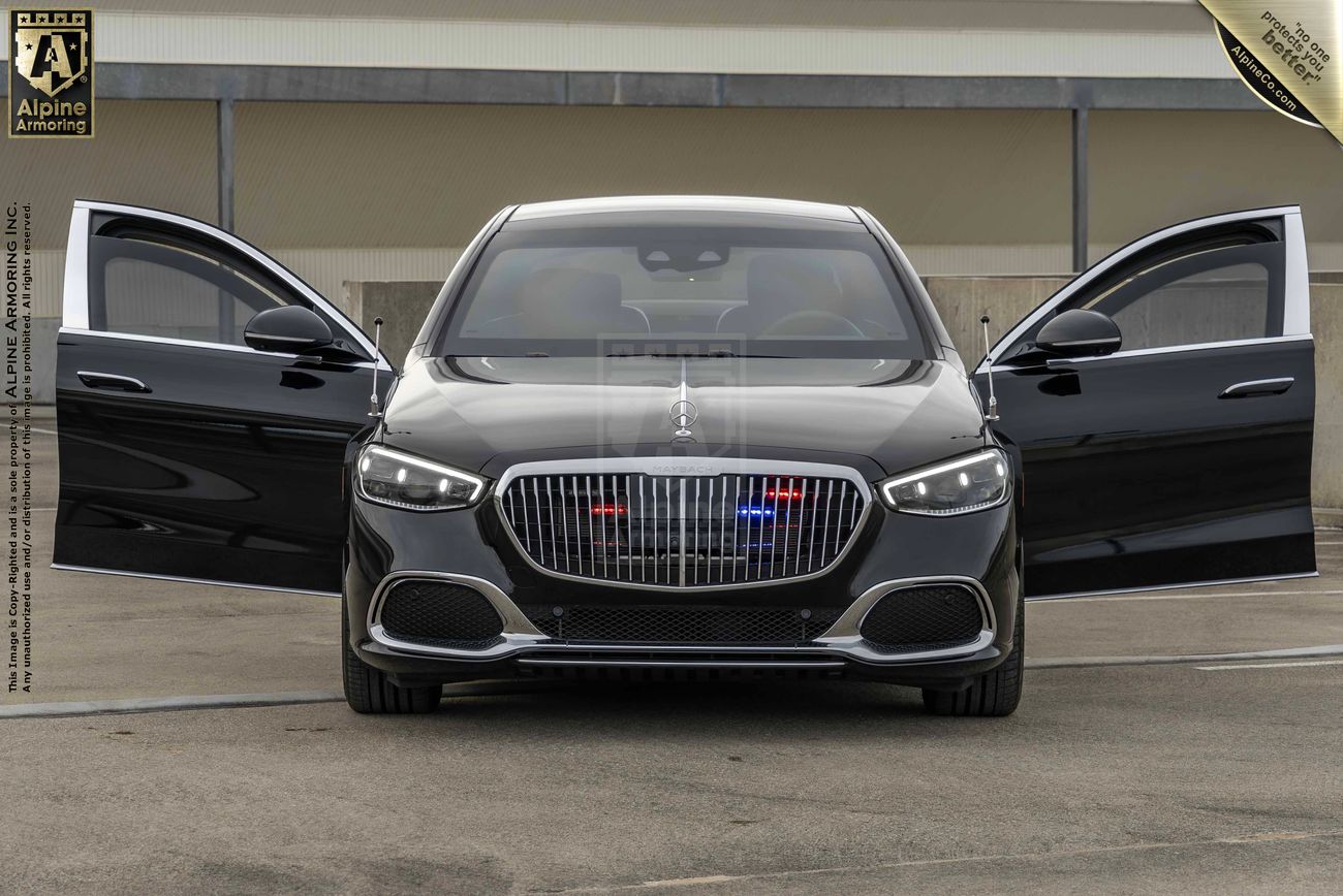 A black armored Mercedes-Benz S 580 Maybach sedan with its front doors open, displaying red and blue lights on the front grill, parked in front of a beige building.