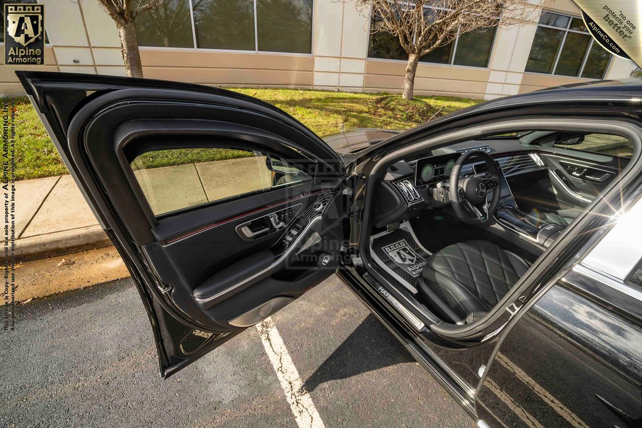 An open driver-side door of a black armored Mercedes Maybach S 580 sedan revealing a clean and well-appointed interior with a sleek dashboard and quilted leather seats. The background shows a building and pavement.