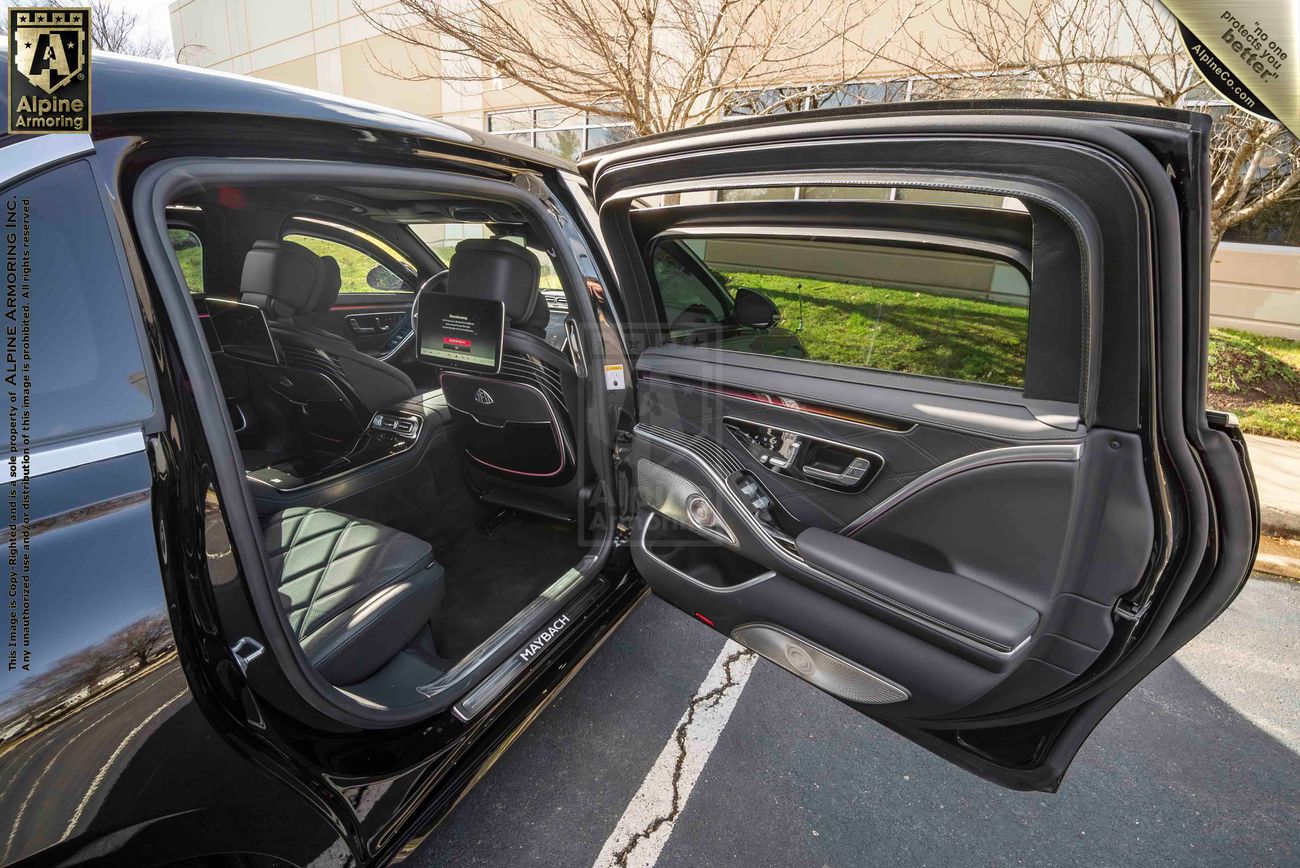 The open door of a black armored Mercedes-Benz S 580 Maybach sedan revealing its spacious back seat with leather upholstery and ample legroom. The car is parked outdoors near a building.
