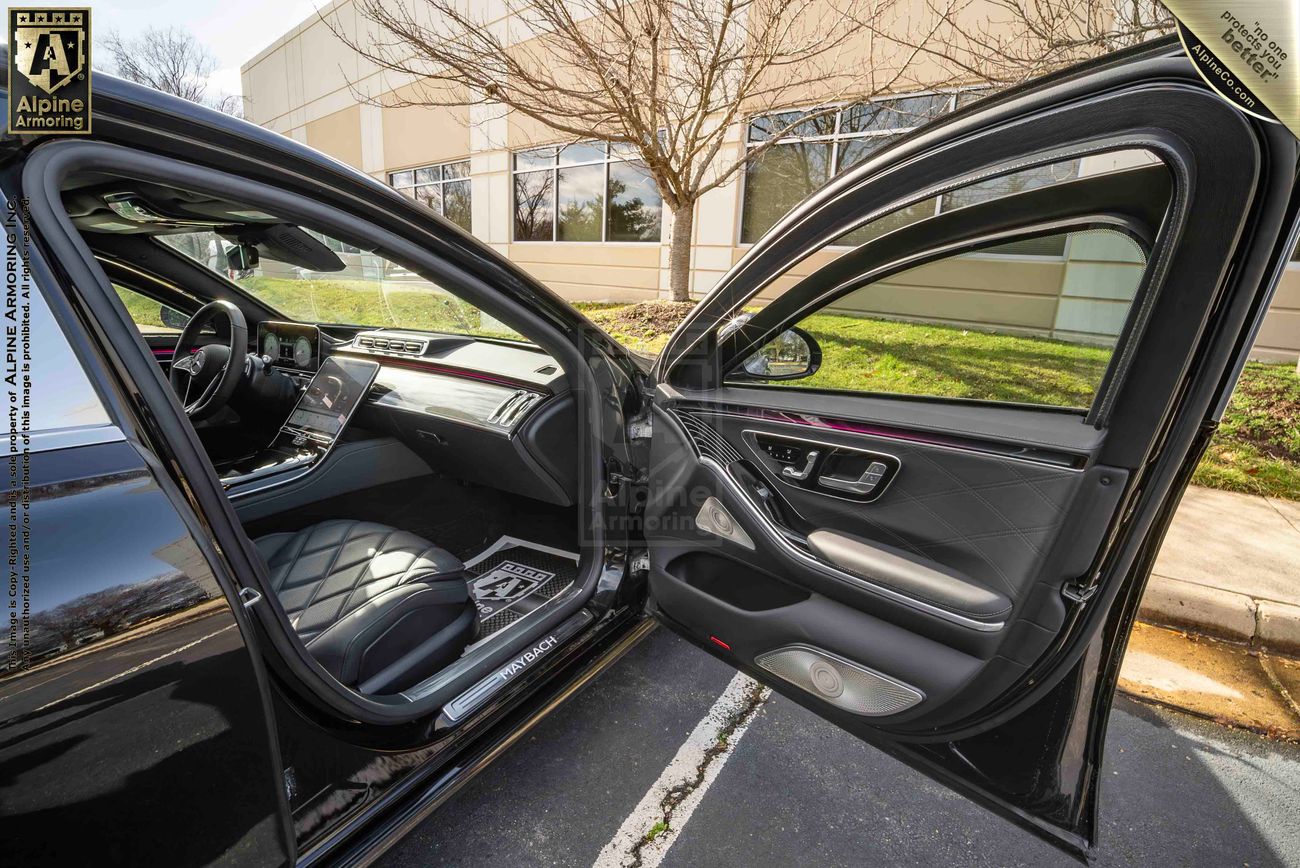 Open passenger-side and rear-side doors of an armored Mercedes-Benz S 580 Maybach sedan showing the interior with leather seats and a modern dashboard. Parked in an outdoor setting near a building.