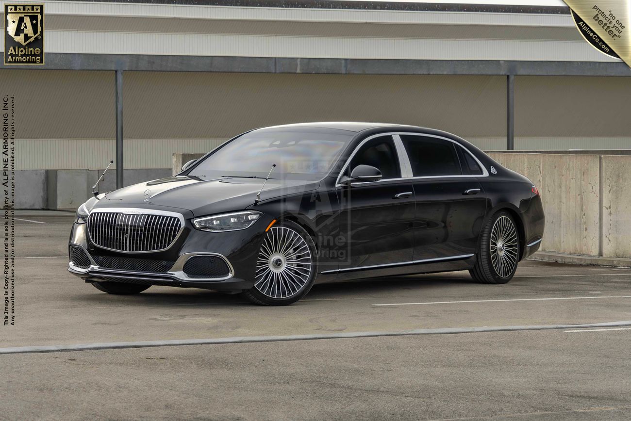 A sleek, black luxury armored Mercedes Maybach S 580 sedan is parked in an outdoor lot against a concrete wall and industrial background.