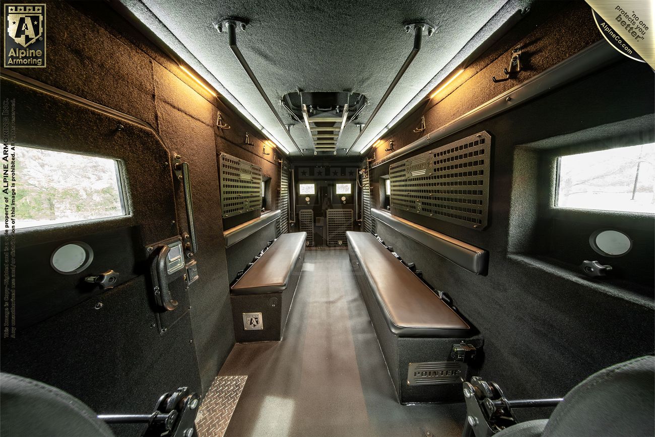 An interior view of a black armored SWAT van, known as the Armored Pointer from Alpine Armoring, featuring reinforced windows, tactical lighting, and advanced security equipment. 