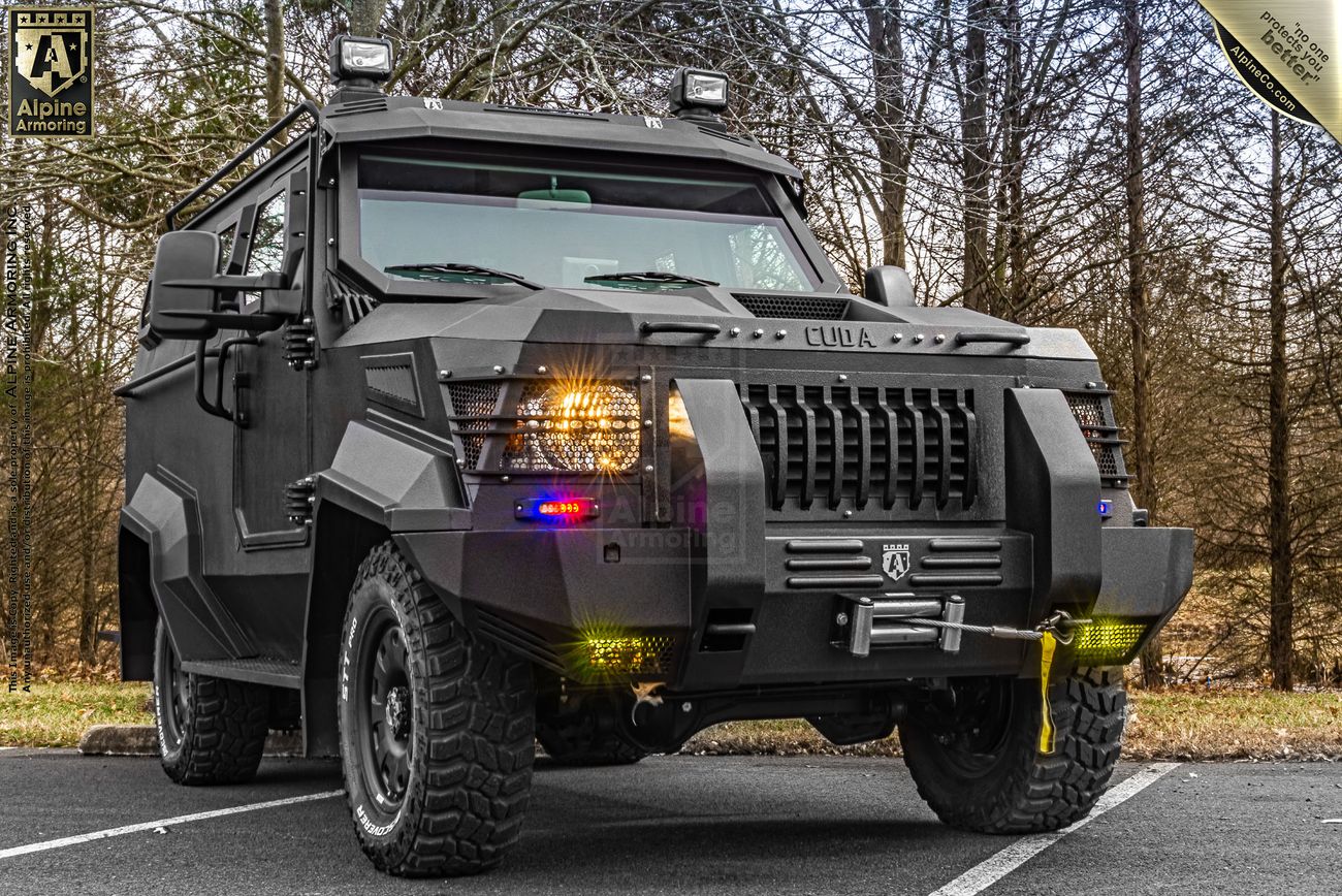 A black armored CUDA is parked on a road with trees in the background. The vehicle is equipped with front lights and rugged tires.