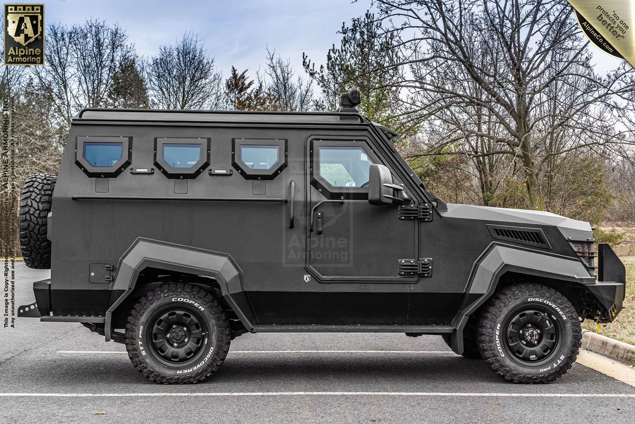A black armored CUDA is parked on a road, featuring multiple small windows and heavy-duty tires. Trees are visible in the background. The vehicle has Alpine Armoring branding.