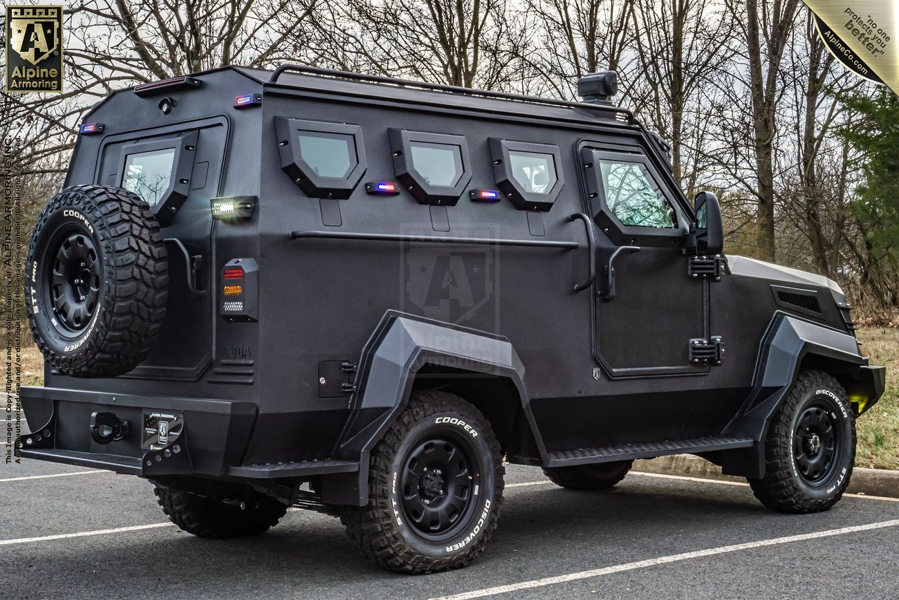 A black armored, bulletproof CUDA with multiple small windows, a mounted spare tire on the back, and rugged tires is parked on a road with bare trees in the background.