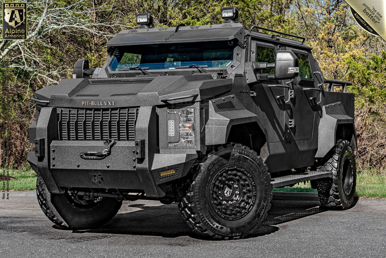 A rugged, armored Pit-Bull VXT APC SWAT truck from Alpine Armoring stands parked on a paved surface, surrounded by trees. The vehicle features heavy-duty tires and a robust exterior designed for protection.