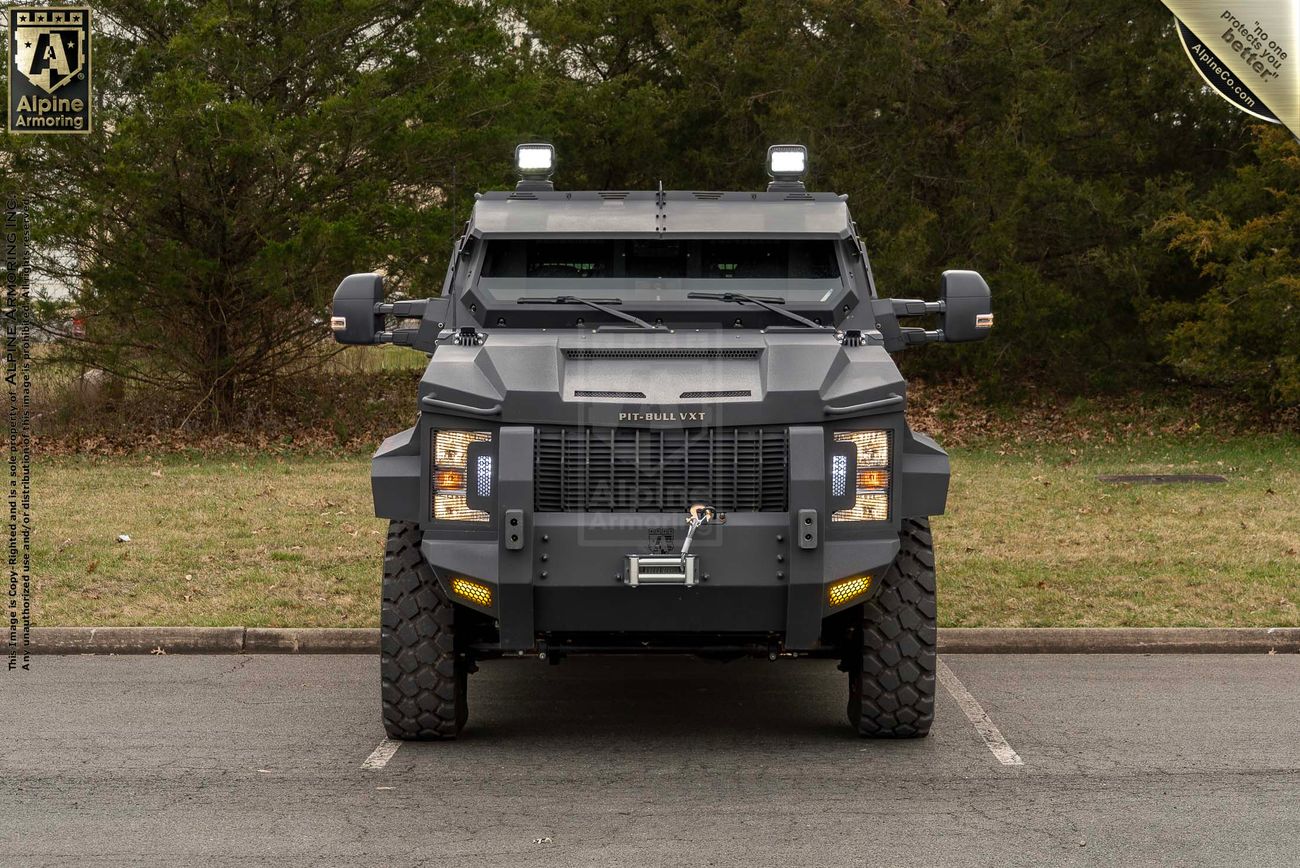 A heavy-duty armored Pit-Bull VXT displayed in a parking lot, seen from the front. The vehicle is equipped with large off-road tires and various protective features. Trees and grass are visible in the background.