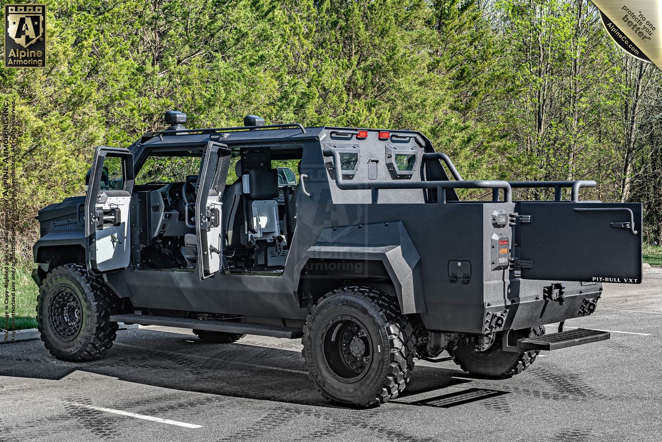 A black armored Pit-Bull VXT with open doors is parked on a road with green trees in the background. The vehicle has rugged tires and a robust design, often used by military or security forces.