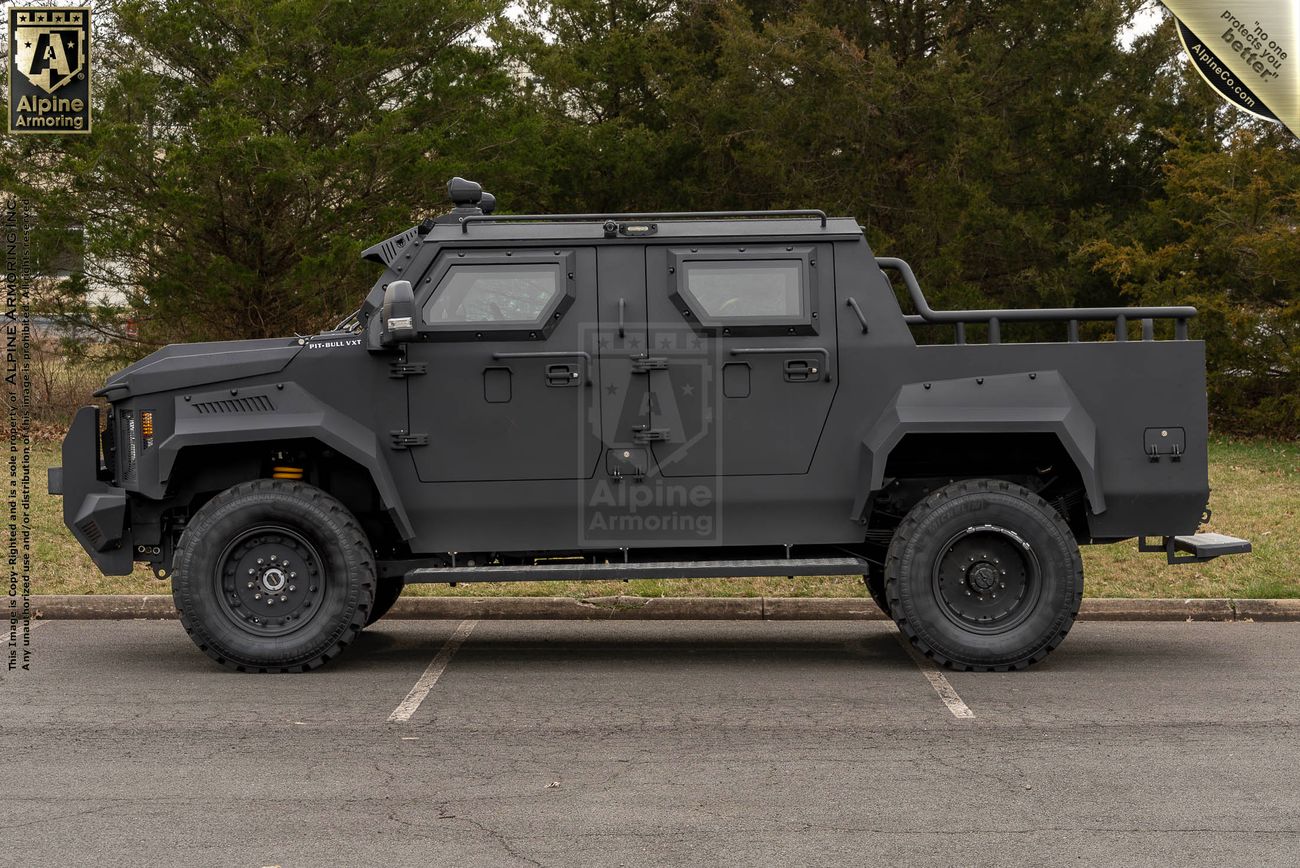 A black armored Pit-Bull VXT is parked in a parking lot. The vehicle has rugged tires, protective plating, and a logo reading "Alpine Armoring" on the side. Trees are visible in the background.