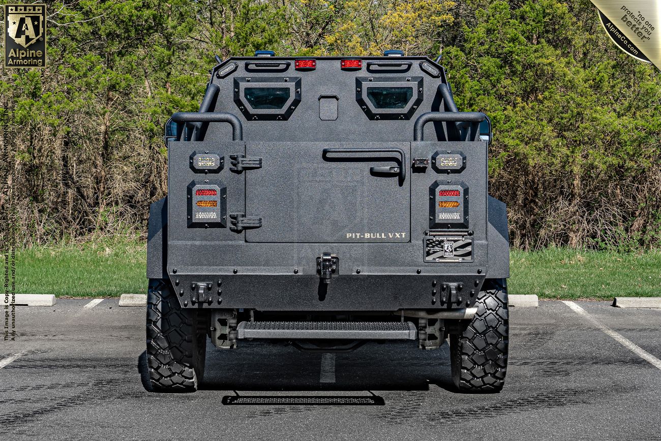Rear view of a Pit-Bull VXT tactical vehicle parked in a lot, showing its rugged design, off-road tires, and various gear and equipment compartments against a backdrop of greenery.