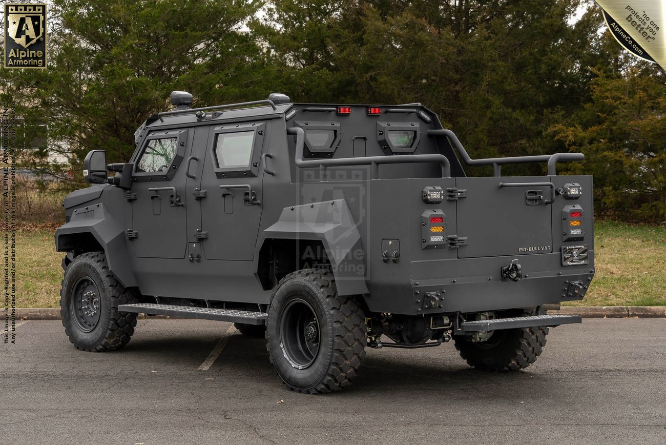 A matte black armored Pit-Bull VXT with rugged tires parked on a paved surface, surrounded by trees.