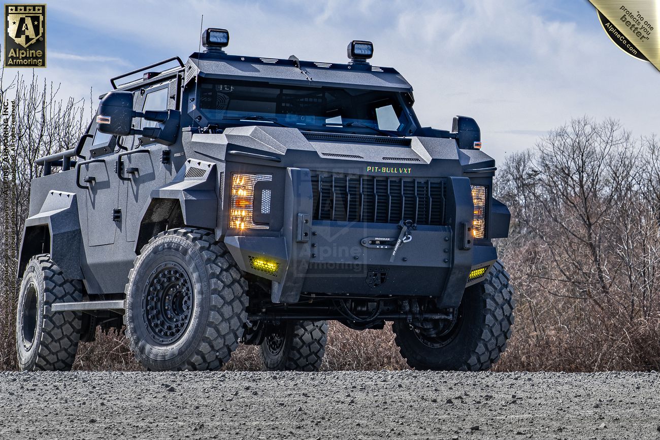 A heavily armored Pit-Bull VXT APC with rugged tires is parked on a gravel surface, featuring prominent front grill and side mirrors, with a cloudy sky backdrop.
