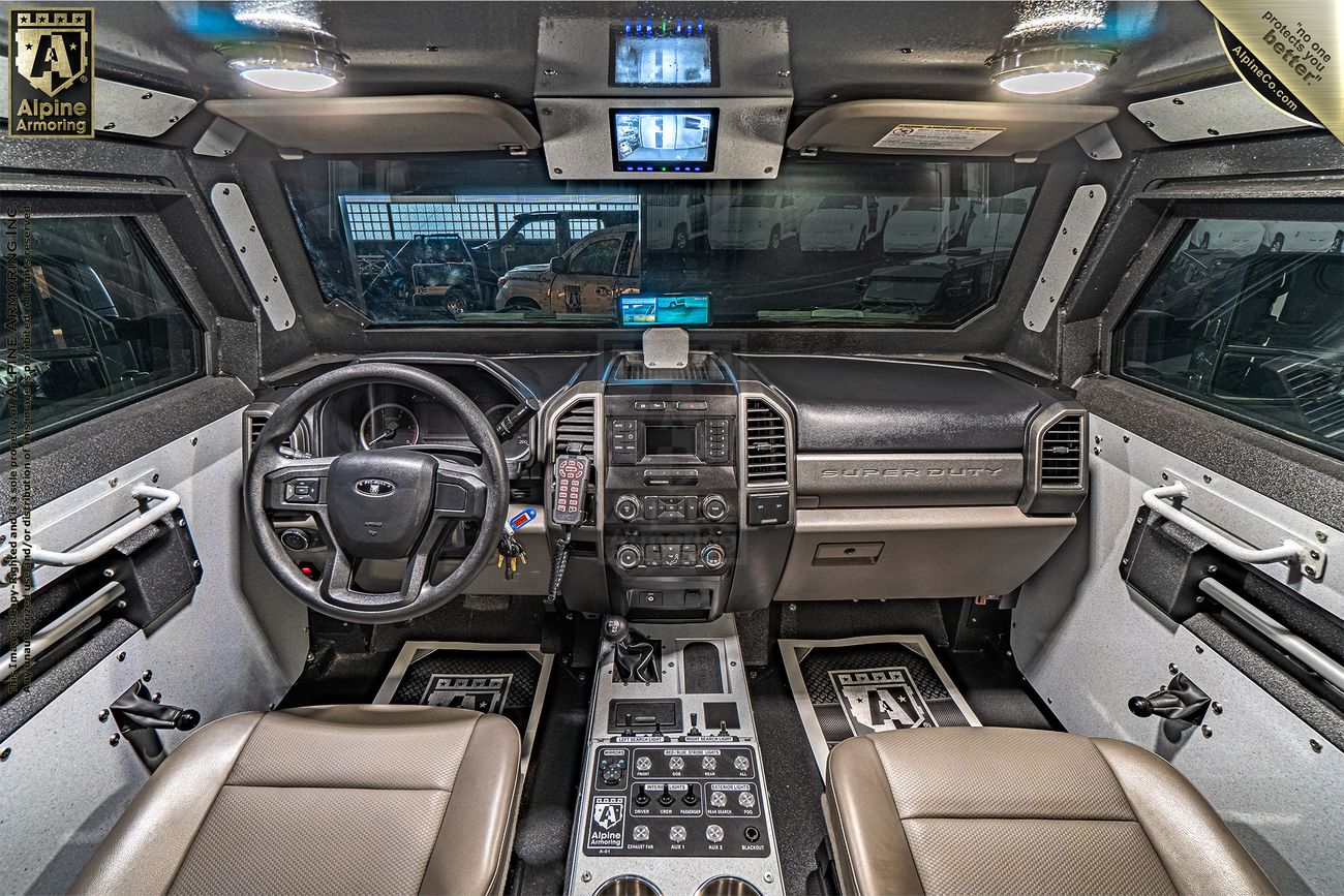 Interior of a Pit-Bull VXT showing a high-tech dashboard with multiple screens, a control panel, and tan leather seats. The Alpine Armoring logo is visible on the upper left side of the image.