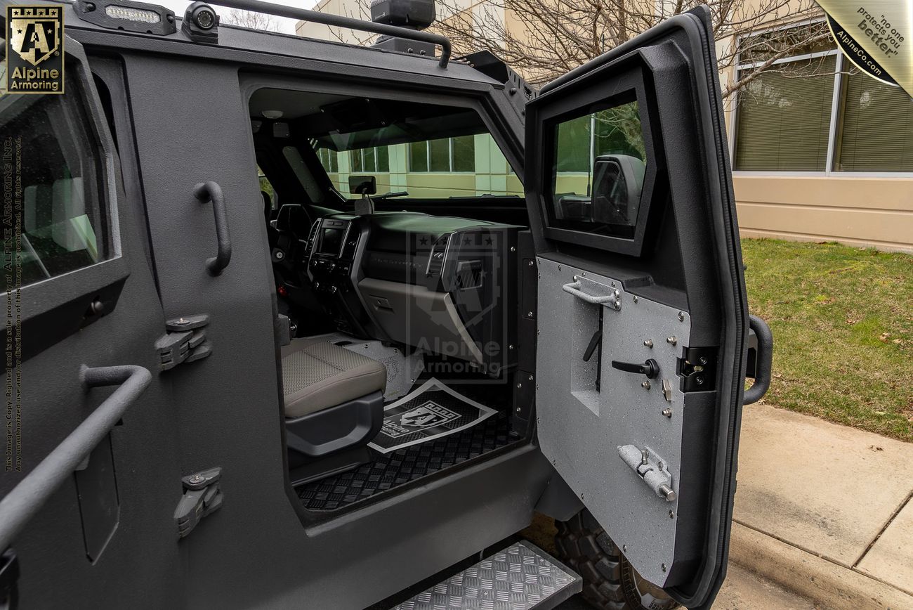 The open passenger door of a bulletproof Pit-Bull VXT, revealing the interior with a step, handle, and seating. The exterior is heavily armored, and a building is visible in the background.