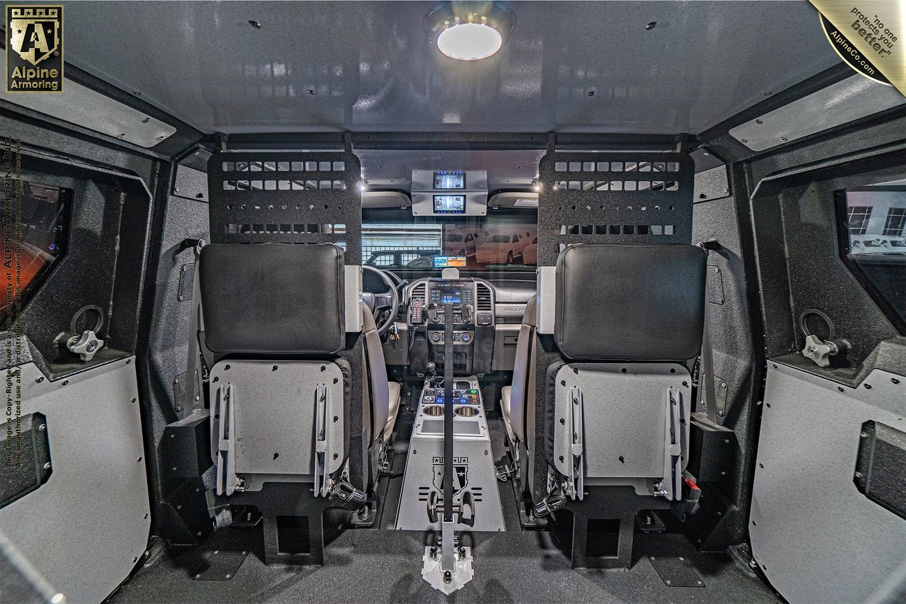 Interior view of an armored Pit-Bull VXT featuring two forward-facing driver and passenger seats, with dashboard and communication equipment visible. The vehicle has reinforced panels and advanced controls.