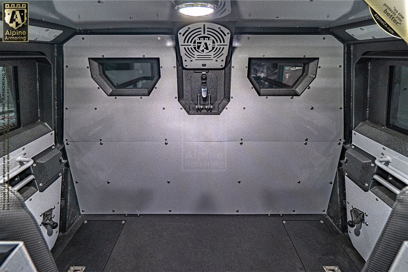 Interior view of a metal-lined vehicle compartment with two small windows and a central ventilation grille. The space is empty with visible bolts and securing mechanisms.