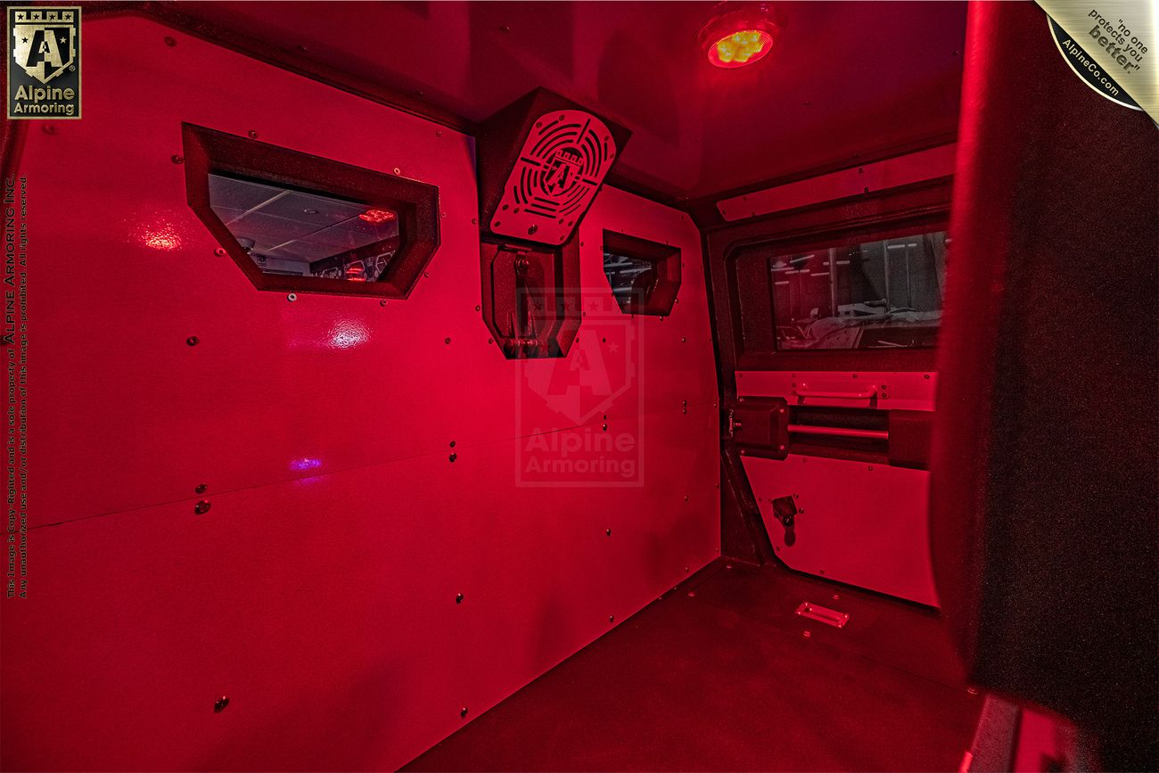 Interior of an armored Pit-Bull VXT with red lighting, featuring reinforced windows and walls, a mounted fan, and a secured compartment. Branding of "Alpine Armoring" is visible on the rear wall.