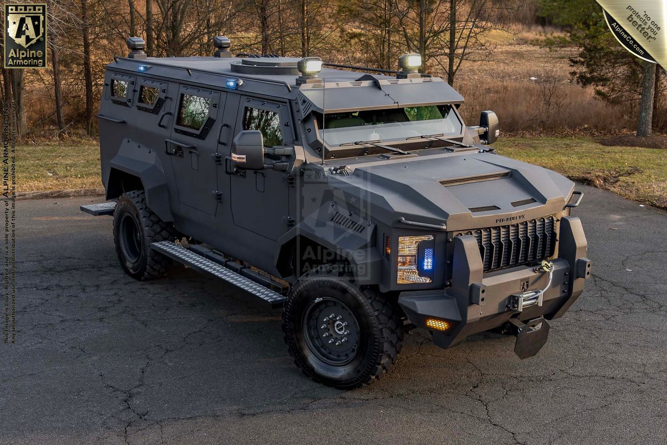 A sturdy, armored Pit-Bull VX 50 cal with rugged tires and various mounted equipment, parked on a road with a grassy background.