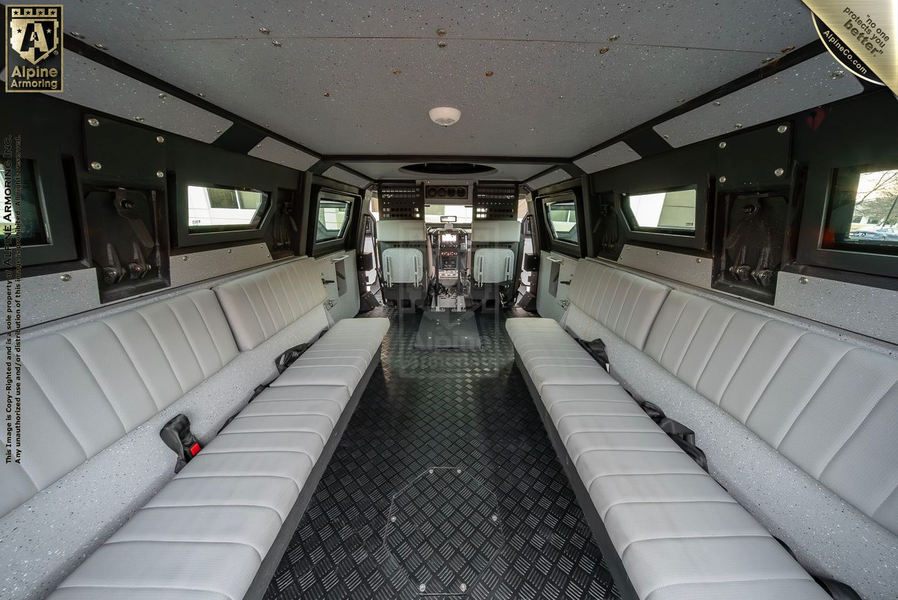 Interior of an armored Pit-Bull VX 50 cal vehicle with two long bench seats along the sides, black diamond-patterned floor, and high ceiling. The driver's cabin with two seats is visible at the front.