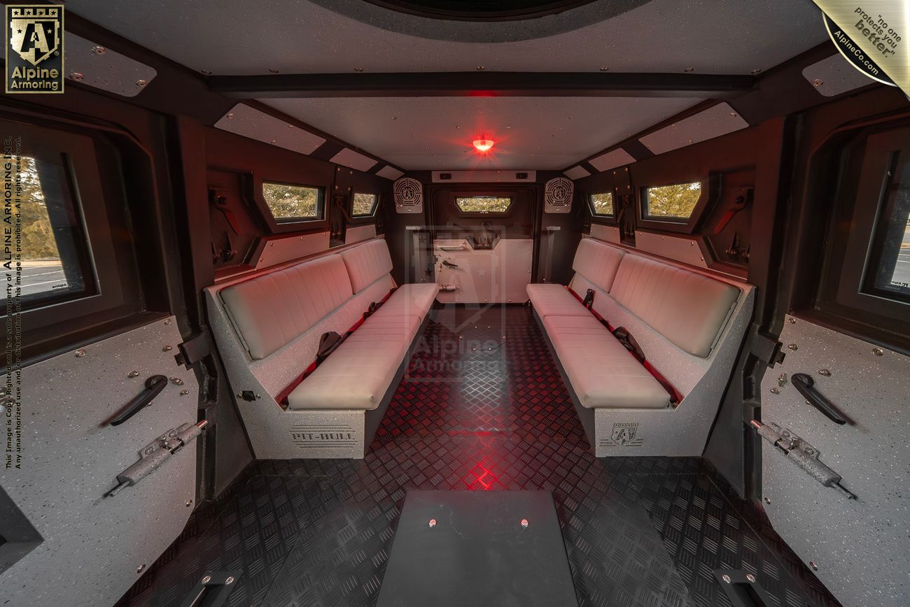 Interior view of an armored Pit-Bull VX 50 cal showing two rows of padded bench seating against the walls, with a red light illuminating the space and small windows on either side.