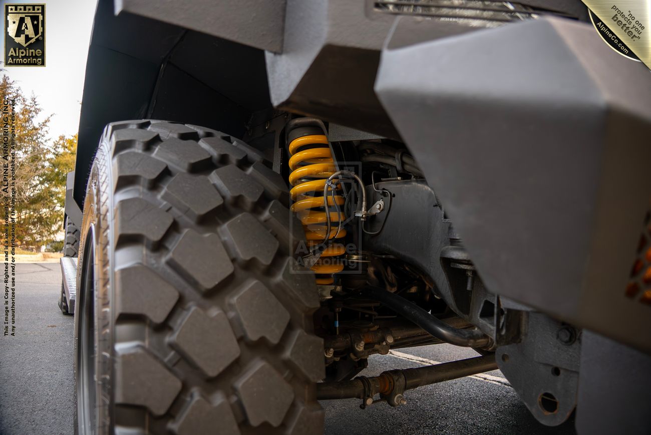 Close-up photo of a rugged Pit-Bull VX 50 cal vehicle suspension system featuring a prominent yellow coil spring and large treaded tire. Logos from Alpine Armoring are visible on the suspension component.