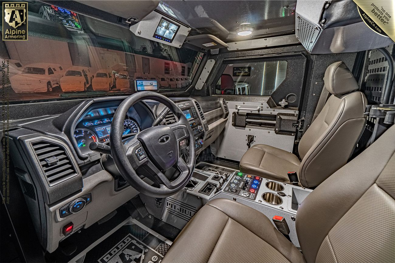 Interior of an armored Pit-Bull VX APC showing the driver's seat, steering wheel, dashboard with gauges, control panels with various buttons, and a monitor screen, all in a highly fortified cabin.