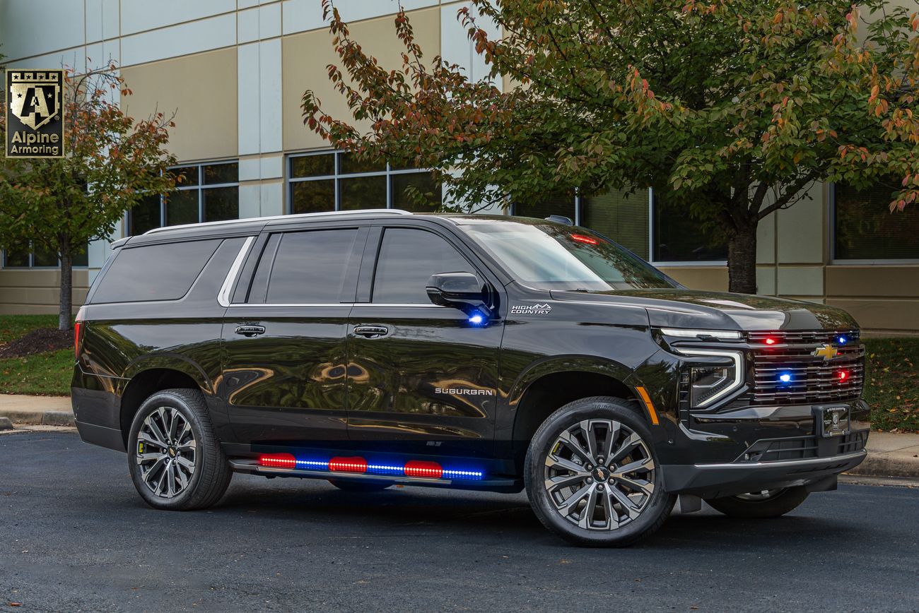 Black armored Chevrolet Suburban High Country SUV with red and blue emergency lights, featuring chrome trim and multi-spoke wheels, parked in front of commercial building